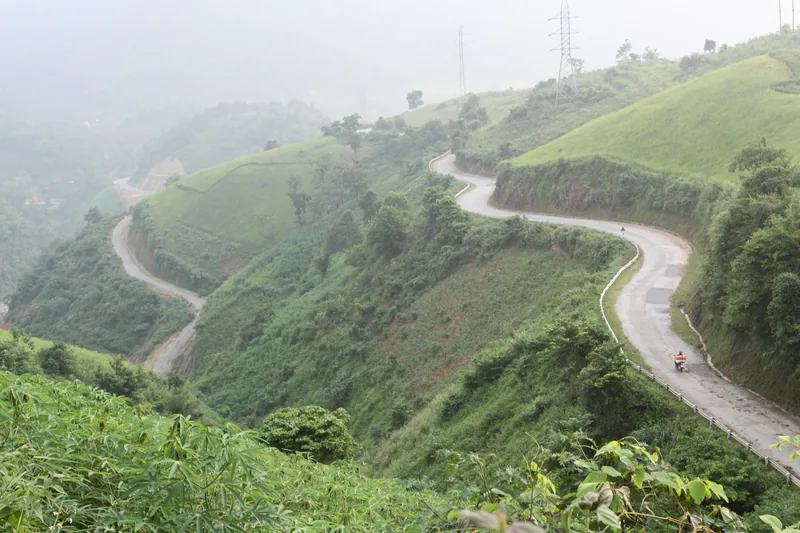 A winding mountain road snakes through green hills shrouded in mist near A Pa Chai, Dien Bien.