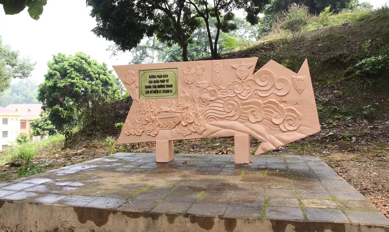 A carved stone monument with relief patterns and an inscription plaque stands on a hillside near Dien Bien.