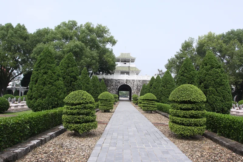 A stone pathway lined with manicured shrubs leads to an arched gate and pagoda-style tower at a war martyrs' cemetery in.