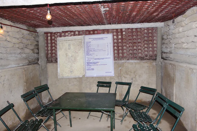 A green metal table and folding chairs sit inside a bunker room with sandbag walls and a battle map at Dien Bien Phu.