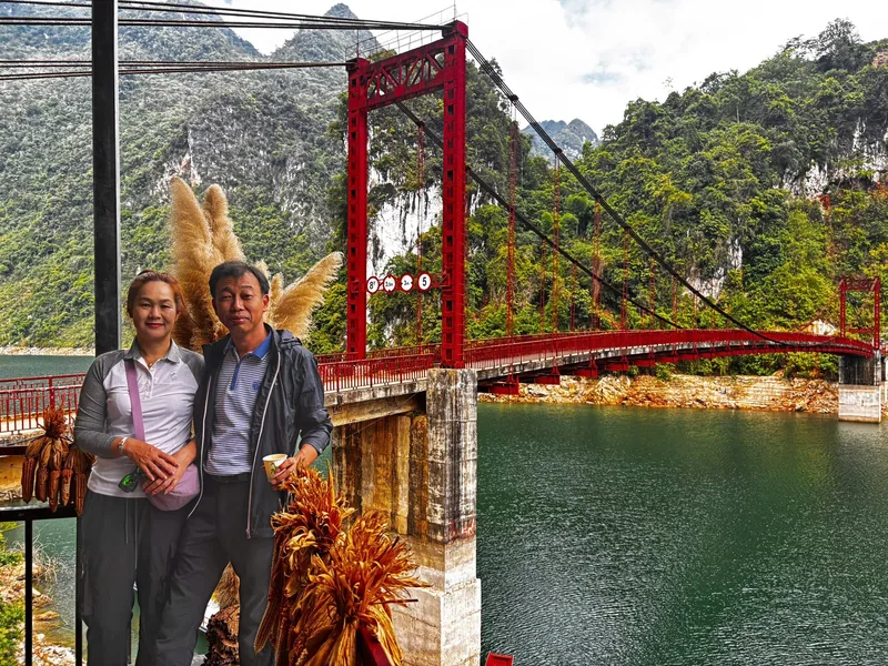A couple poses beside a red suspension bridge over a green reservoir surrounded by limestone mountains near Dien Bien Phu.