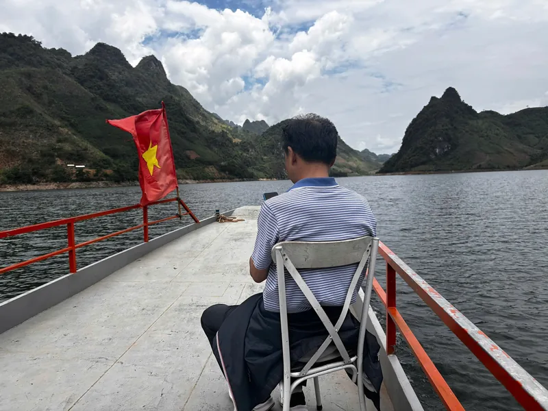 A man sits on a boat deck with a Vietnamese flag, gliding past limestone karst mountains on a reservoir in Dien Bien Phu.