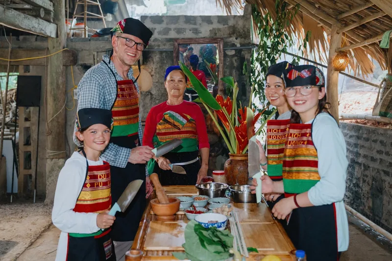 A family in traditional aprons and hats joins a local woman for a cooking activity in a rustic kitchen in Dien Bien.