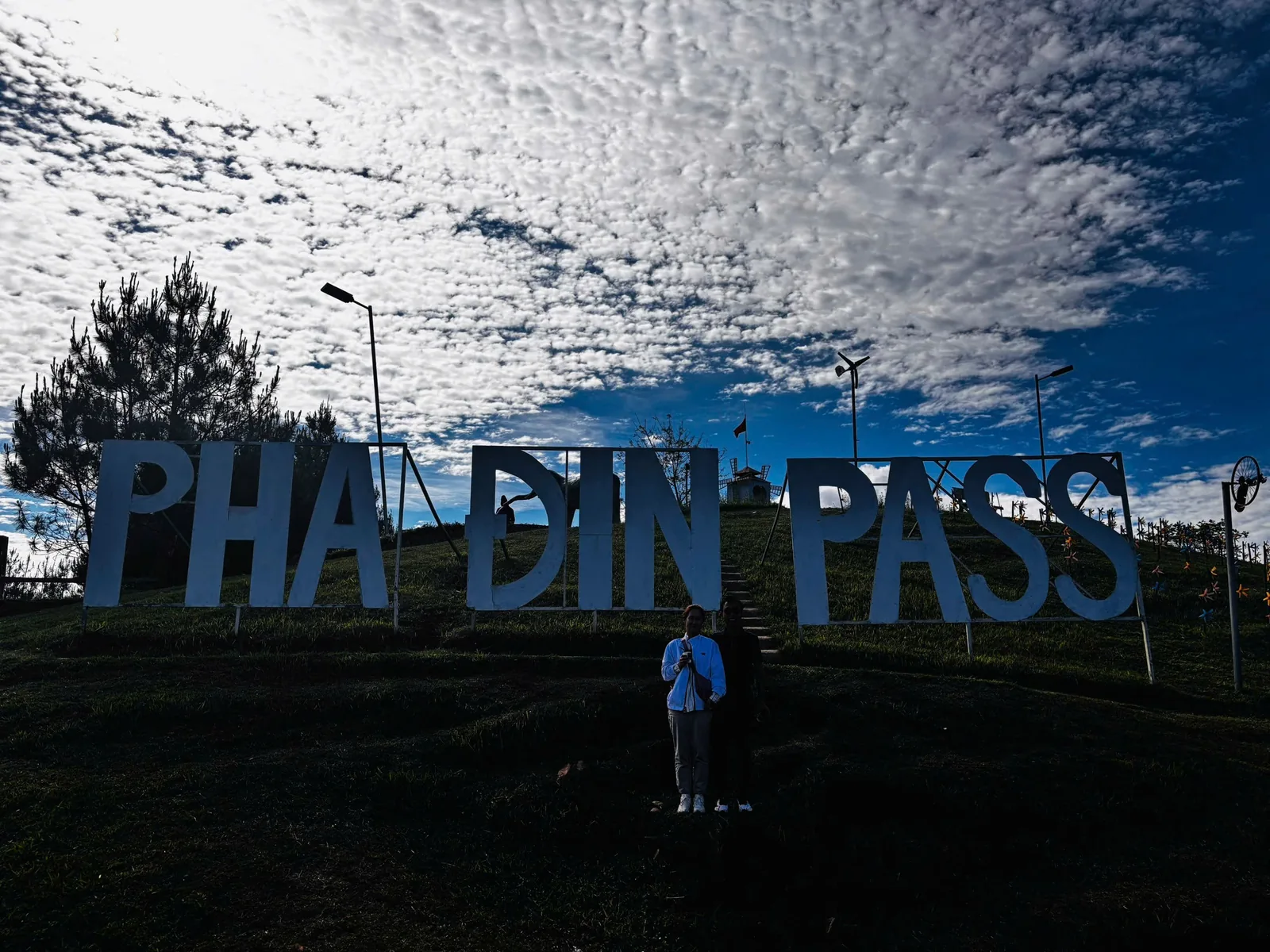 Large letter sign reading Pha Din Pass stands on a hilltop under a dramatic sky with clouds and blue sky.
