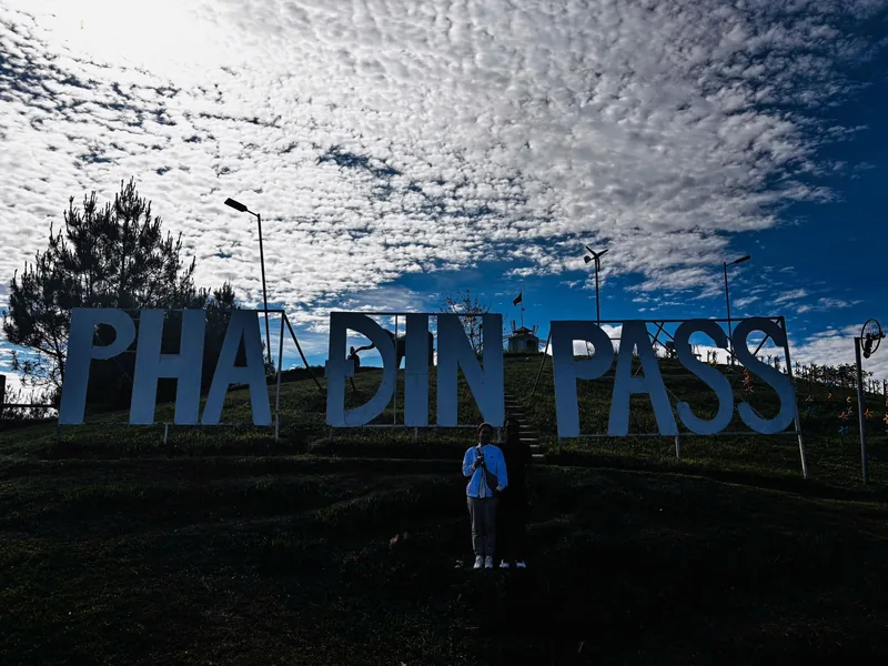 Large letter sign reading Pha Din Pass stands on a hilltop under a dramatic sky with clouds and blue sky.