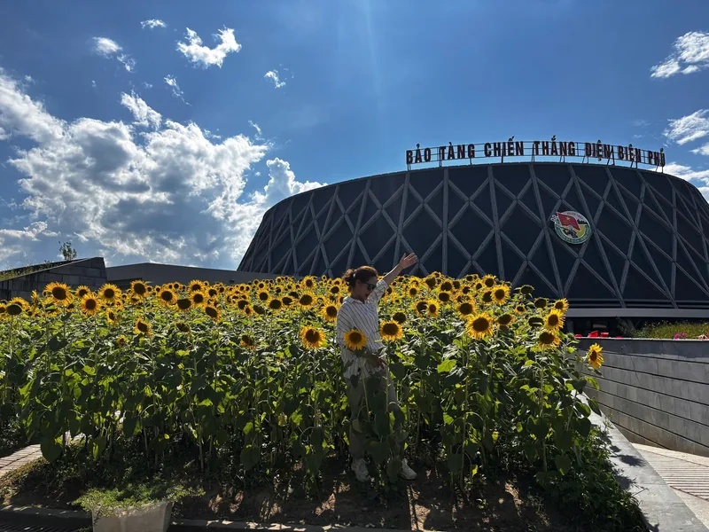A woman poses among sunflowers in front of the Dien Bien Phu Victory Museum under a bright blue sky.