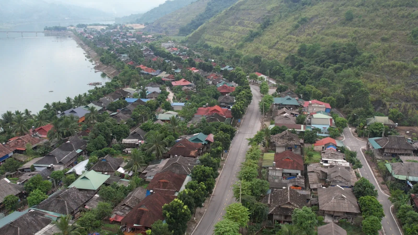 Aerial view of a riverside village in Dien Bien with a road running through rooftops between hills and water.